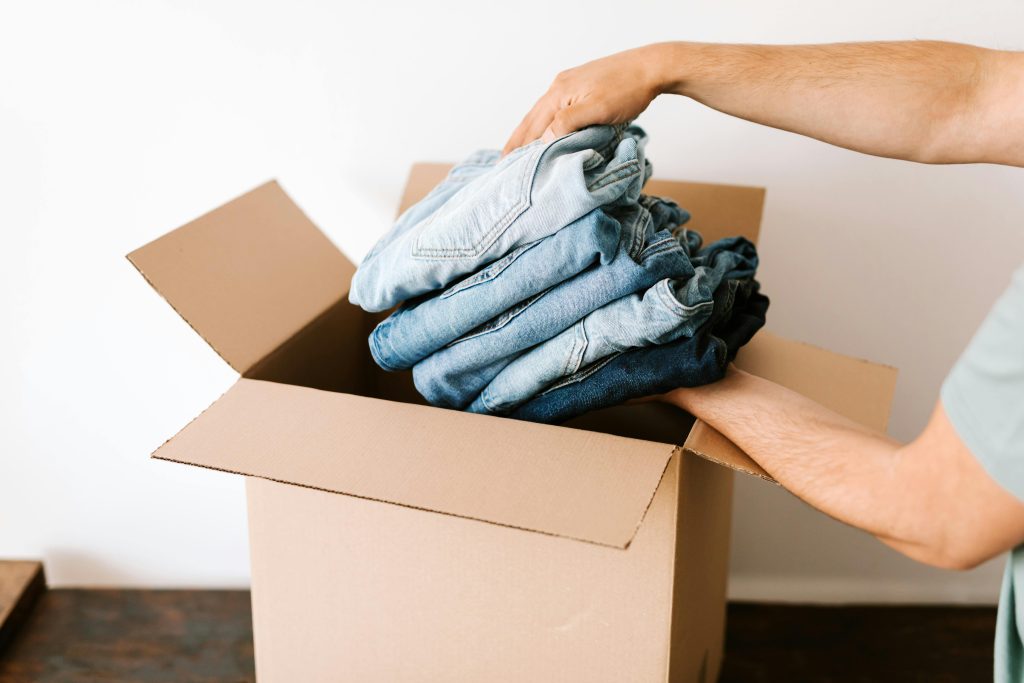 Crop unrecognizable male putting staked jeans into opened cardboard container before renovation while standing against white wall in daylight