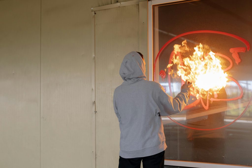 Person in gray hoodie using spray can on glass with flames visible, suggesting vandalism.