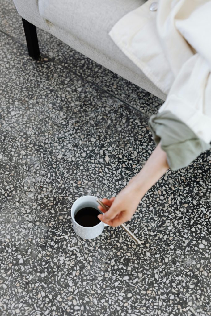 A person stirs coffee in a ceramic cup while seated on a terrazzo floor indoors.